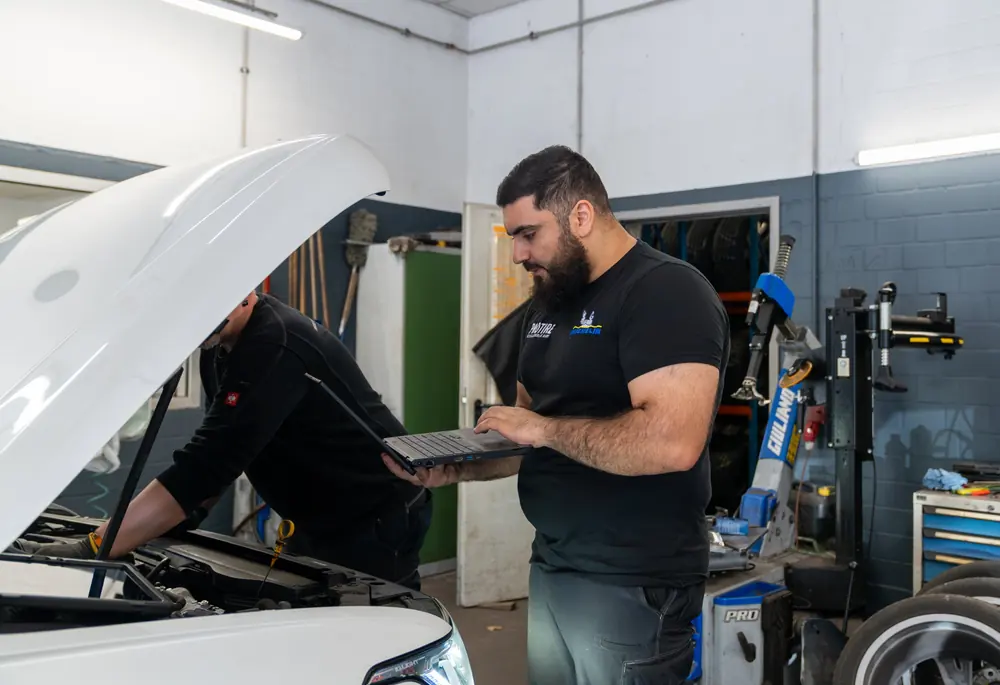 Zwei Mechaniker bei der Fahrzeugdiagnose mit Laptop am offenen Motorraum